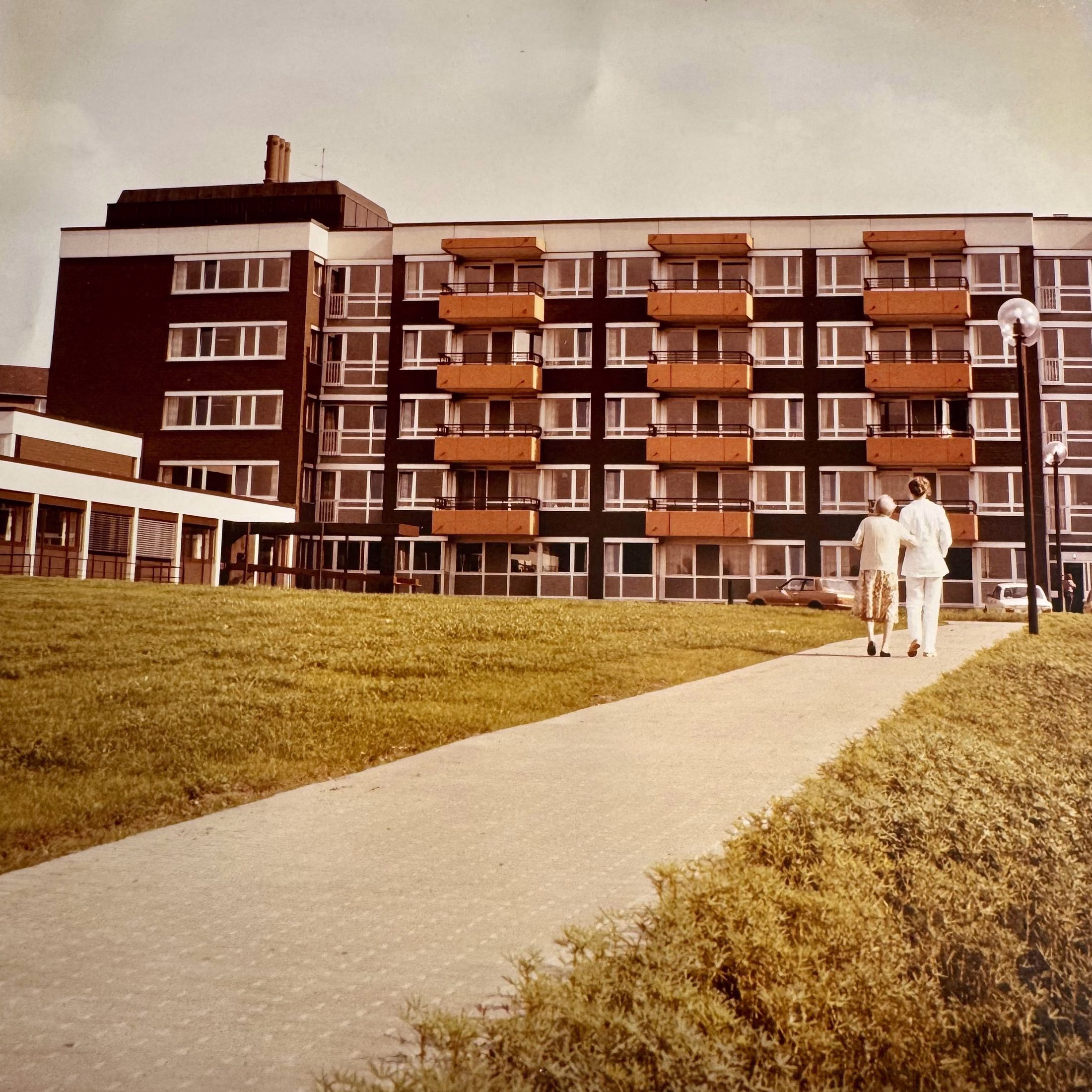 Eine Pflegekraft in Weiß und eine ältere Dame gehen von hinten gesehen Seite an Seite auf einem Weg zum Haus Auf dem Bruch.