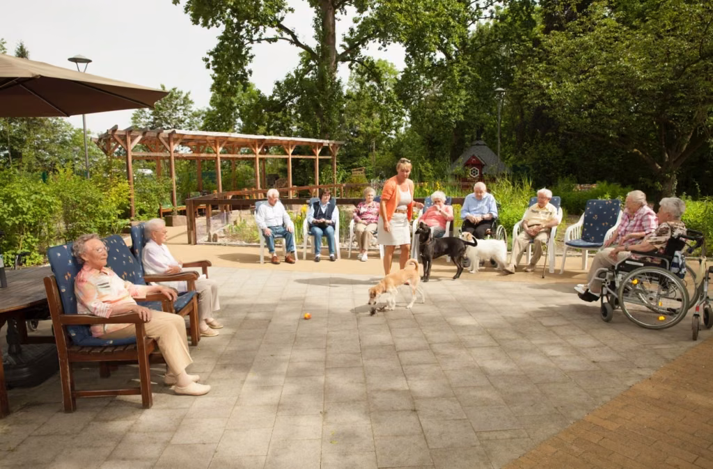 Eine Gruppe sitzt im Garten im Halbkreis; in der Mitte stehen eine Frau und mehrere Hunde.