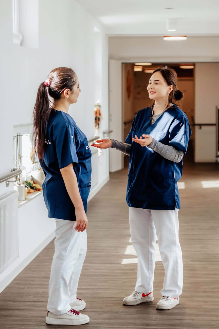 Zwei Frauen in Arbeitskleidung stehen im Flur am Fenster und unterhalten sich.