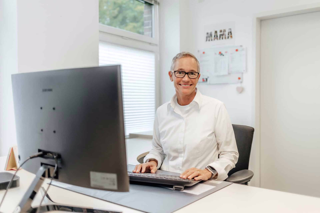 Eine Frau sitzt lächelnd an einem Schreibtisch mit Bildschirm und Tastatur.