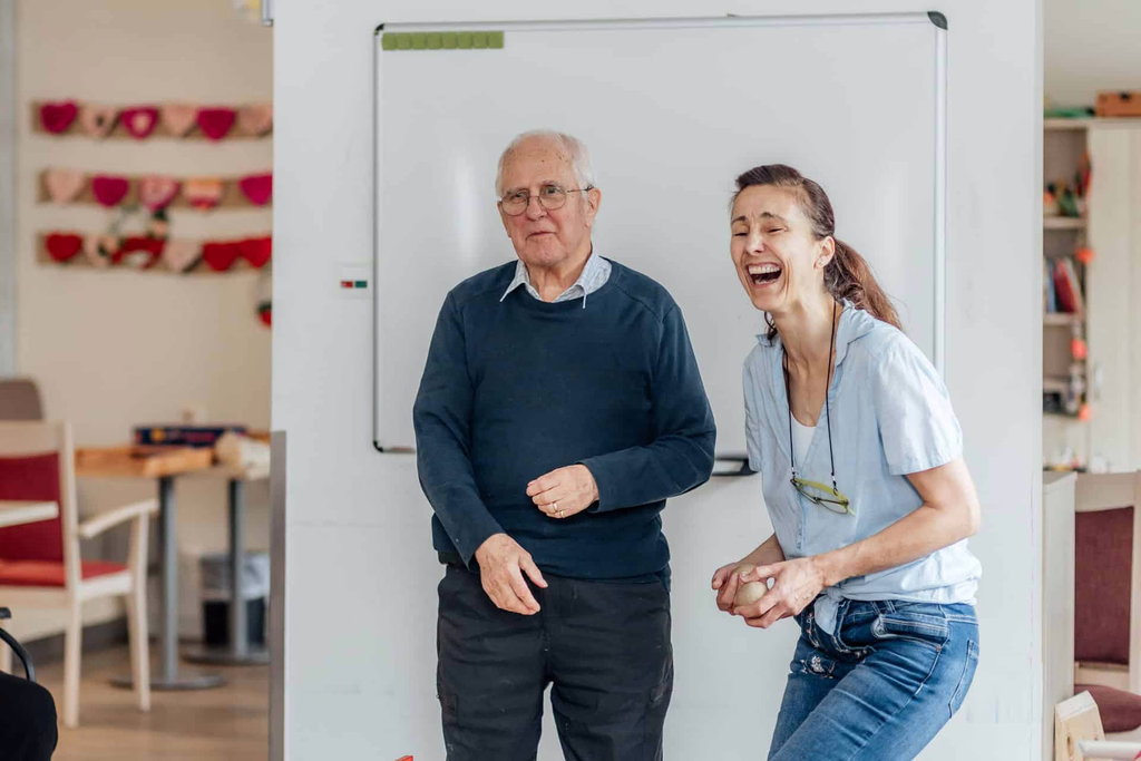 Ein älterer Mann und eine Frau lachen nebeneinander in einem hellen Gemeinschaftsraum.