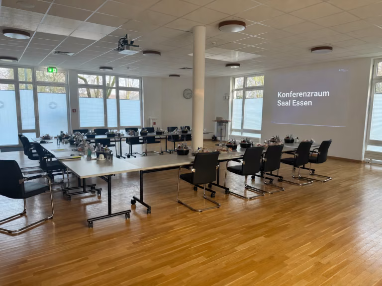 Vorbereiteter Konferenzraum mit Tischen, Stühlen und der Wandprojektion „Konferenzraum Saal Essen“.